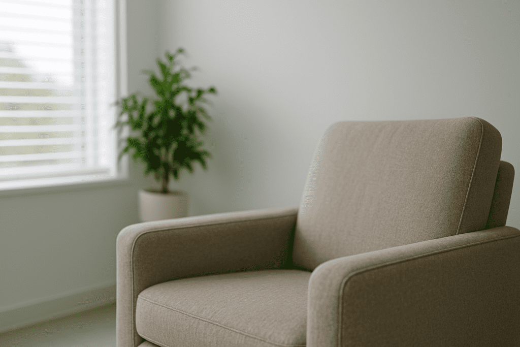 A beige upholstered armchair sits near a large window with white blinds, surrounded by soft natural light and a green potted plant in a modern, minimalist room.